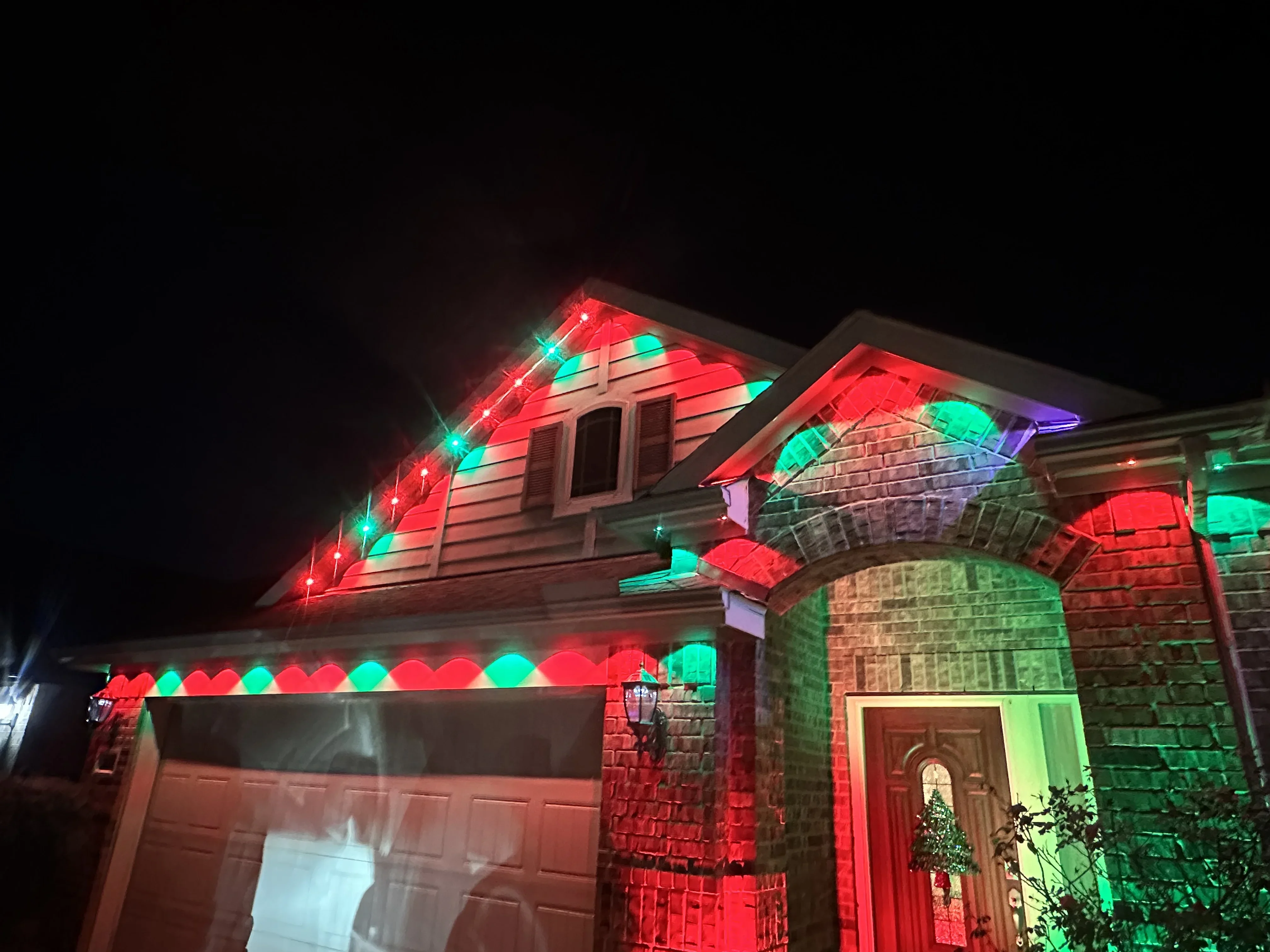 Red and cyan LED holiday lights installed on brick home roofline, gables, and front entry archway at night