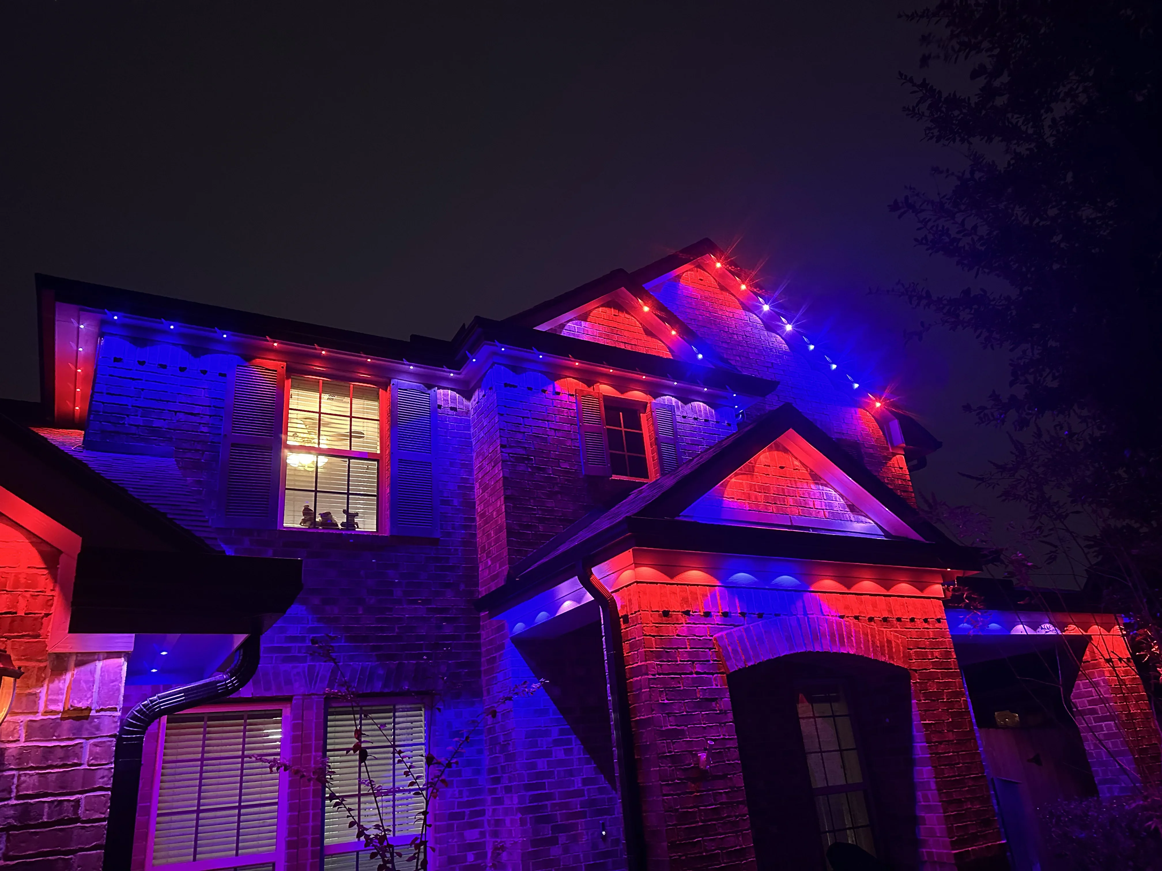 Two-story brick home illuminated with blue and red RGB LED lighting on rooflines and exterior walls at night