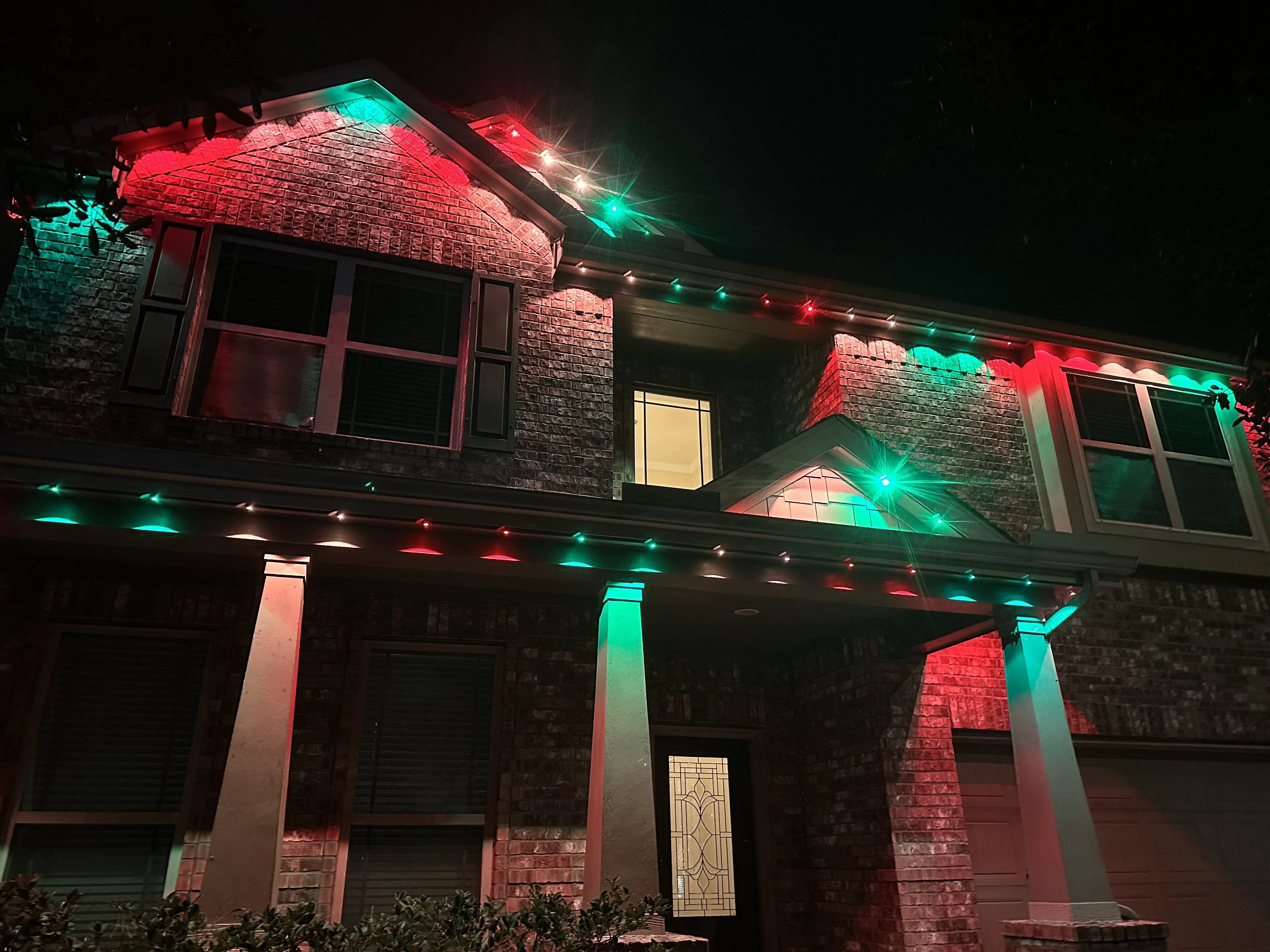 Two-story brick home with red, green, and white LED holiday lights on roofline and columns