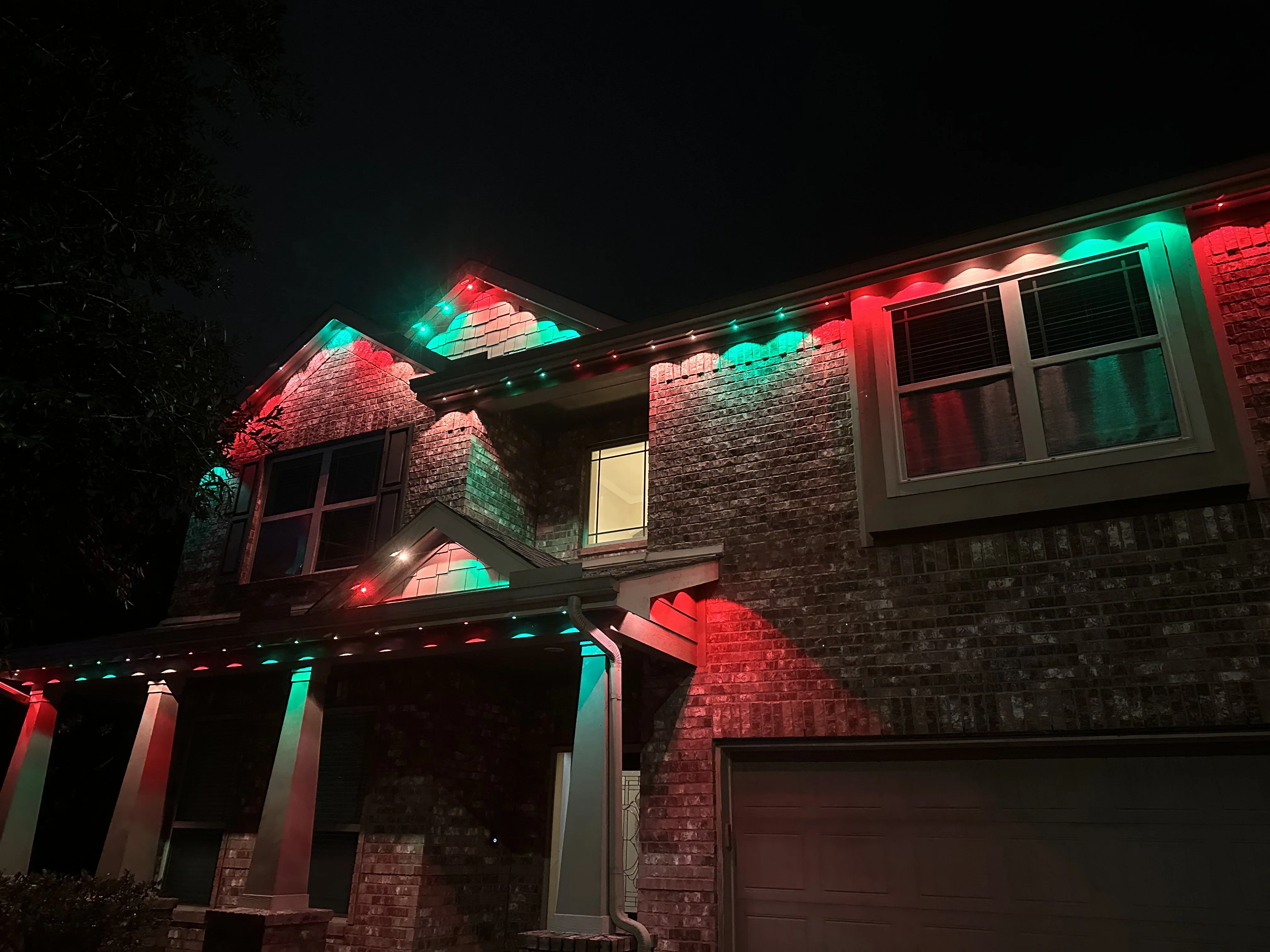 Two-story brick home with red and cyan LED accent lighting on roofline, gables, and columns at night
